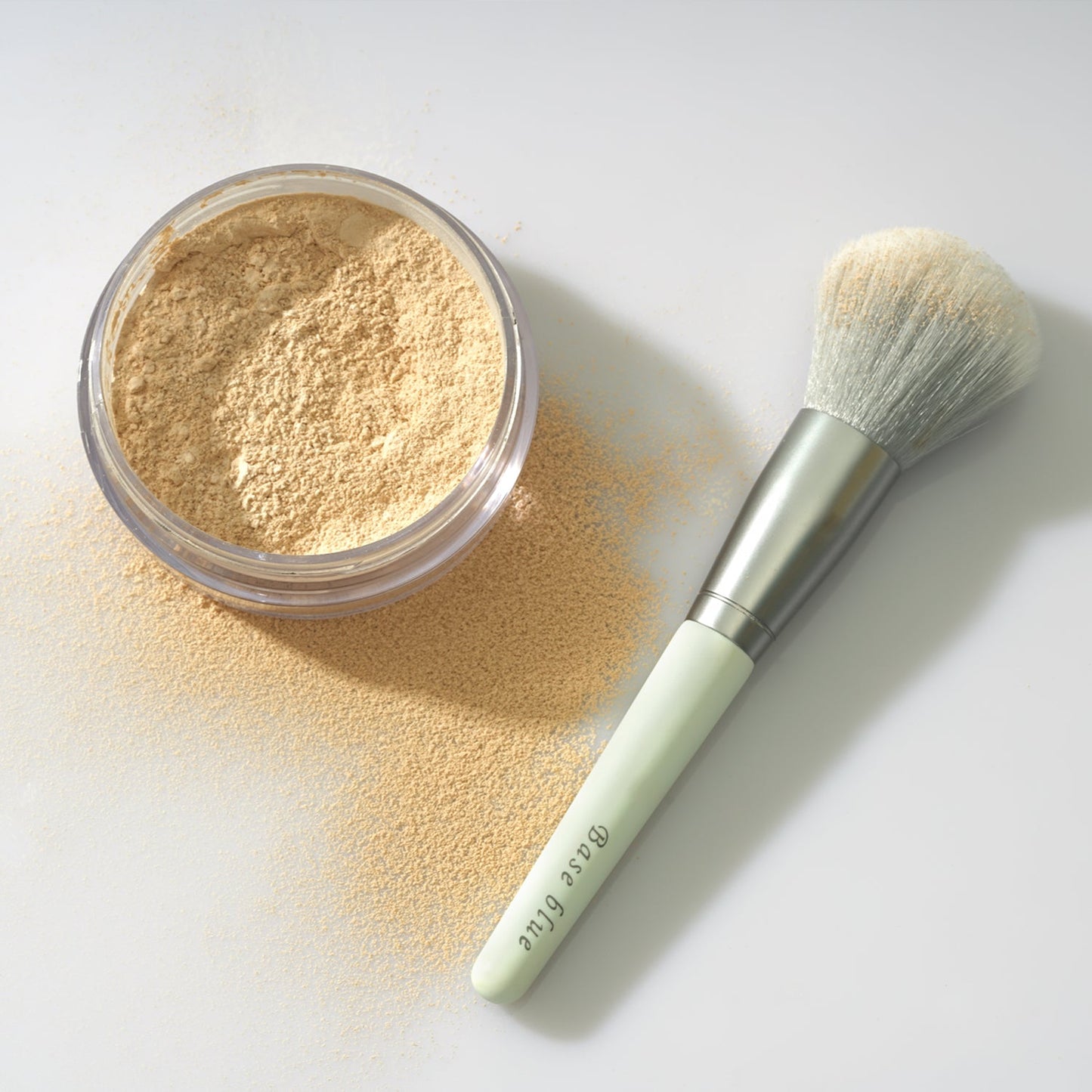 Image of Baseblue Mini Soft Brush next to a jar of powder, a compact travel-sized makeup brush for on-the-go beauty routines.